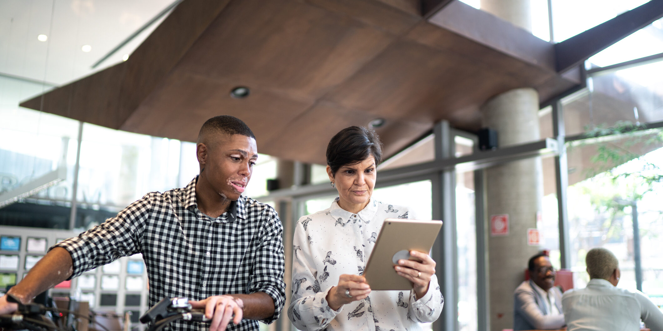 Coworkers talking in office lobby, carrying a bike and showing something in a digital tablet